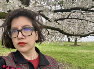 A women who is wearing glasses with chin-length brown hair takes up the left side of the frame. A white cherry blossom tree is behind her.